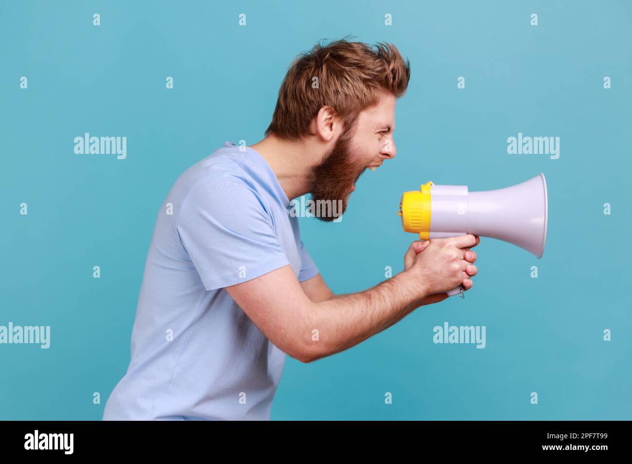 Side view of bearded man holding megaphone near mouth, loudly speaking, screaming, making announcement, paying attention at social problems. Indoor studio shot isolated on blue background. Stock Photo