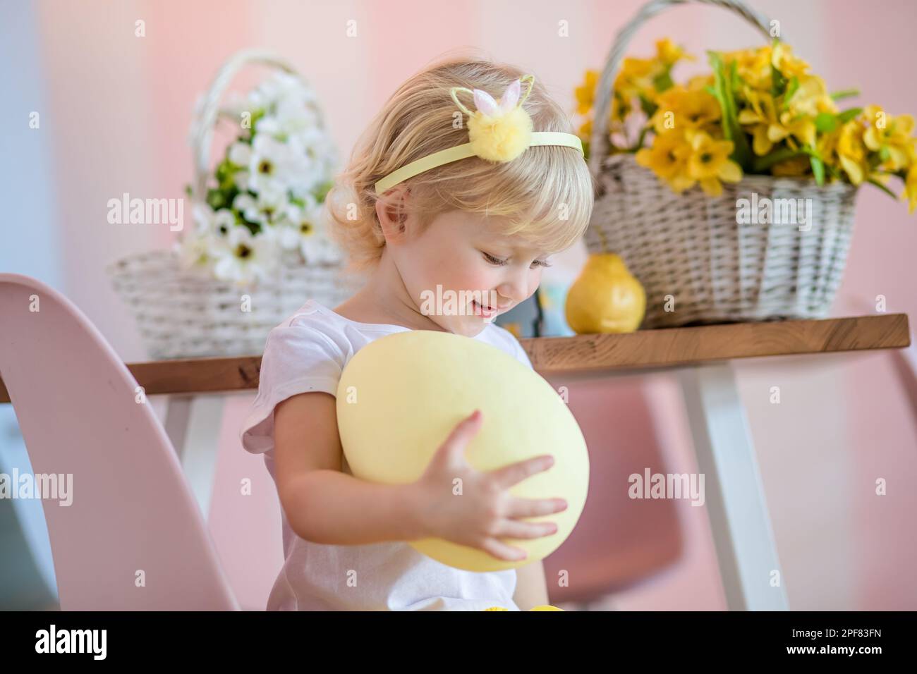 Easter egg hunt at home. Adorable happy little blonde girl with short hair in a white dress holding a huge painted Easter egg Stock Photo