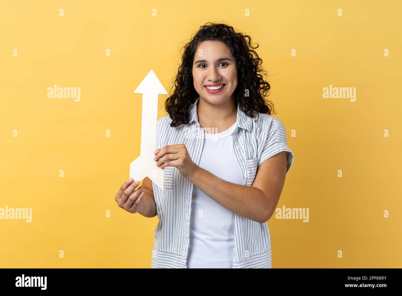Portrait of smiling satisfied woman with dark wavy hair pointing up looking at camera with smile, growth and increase concept. Indoor studio shot isolated on yellow background. Stock Photo
