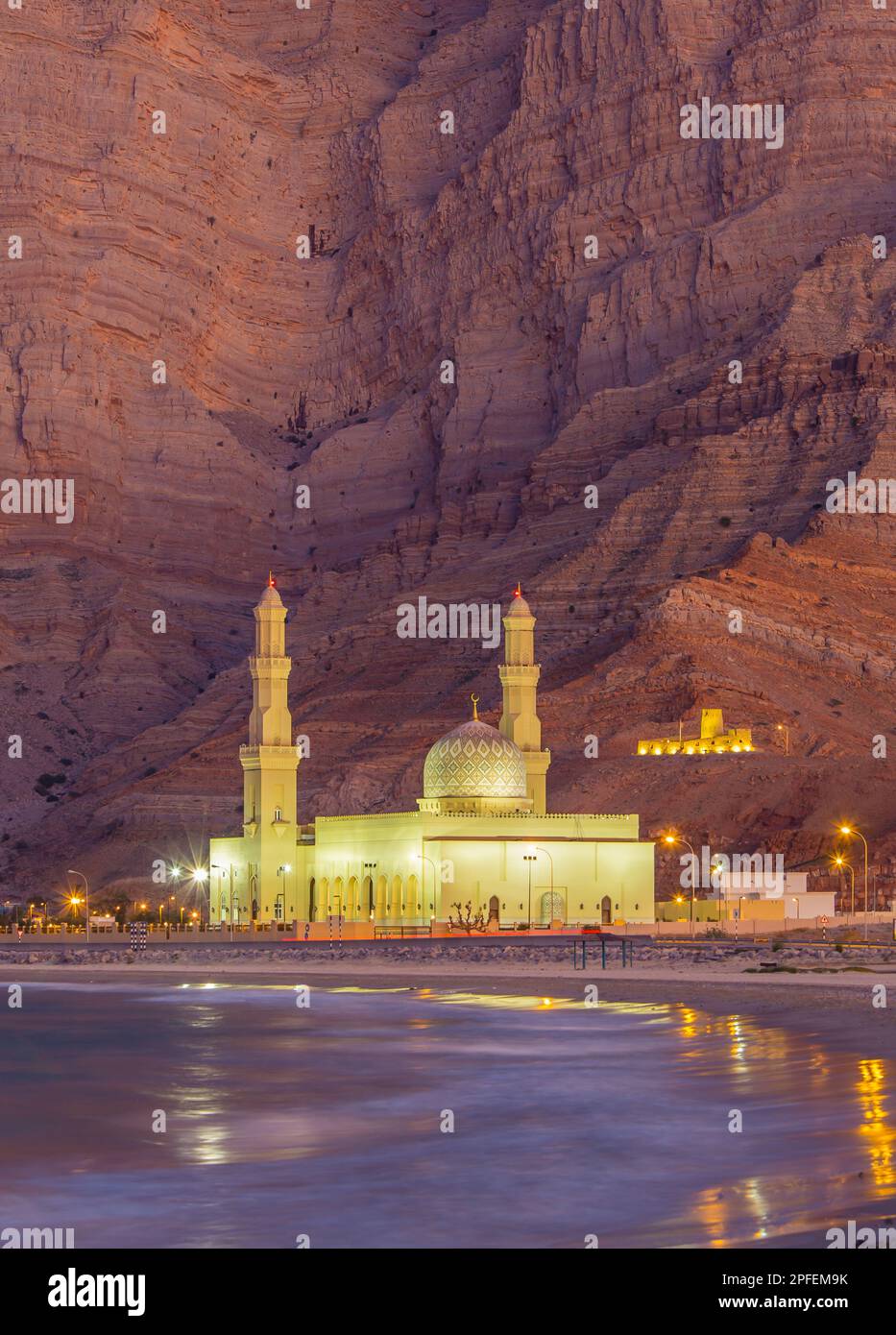 Dramatic rocky backdrop behind the illuminated Khasab Mosque at blue hour in Khasab, Oman Stock Photo