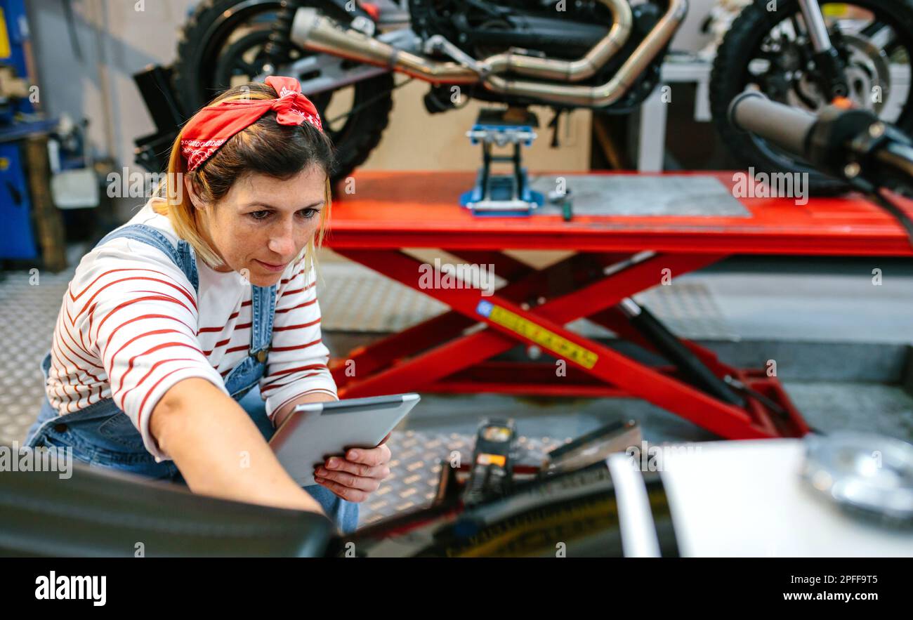 Concentrated female mechanic with digital tablet reviewing information of motorcycle on garage Stock Photo