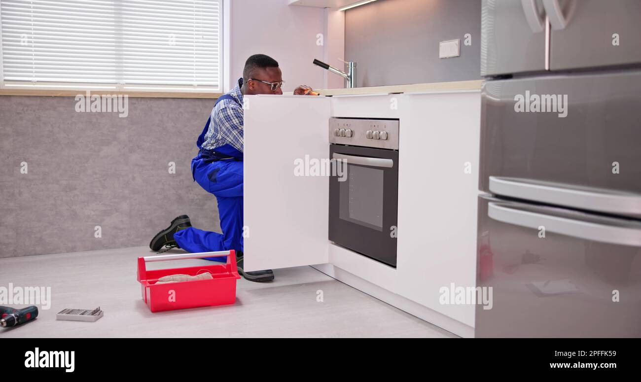 Repairman In Overalls Repairing Cabinet Hinge In Kitchen Stock Photo