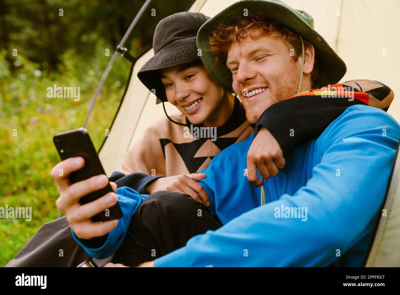 Young cheerful multiethnic tourist couple wearing panama hats taking selfie while sitting at camping tent in green forest Stock Photo