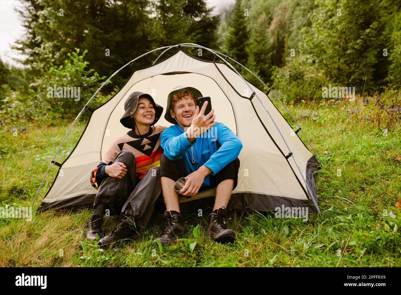Joyful multiethnic tourist couple wearing panama hats taking selfie while  sitting at camping tent in green forest Stock Photo - Alamy