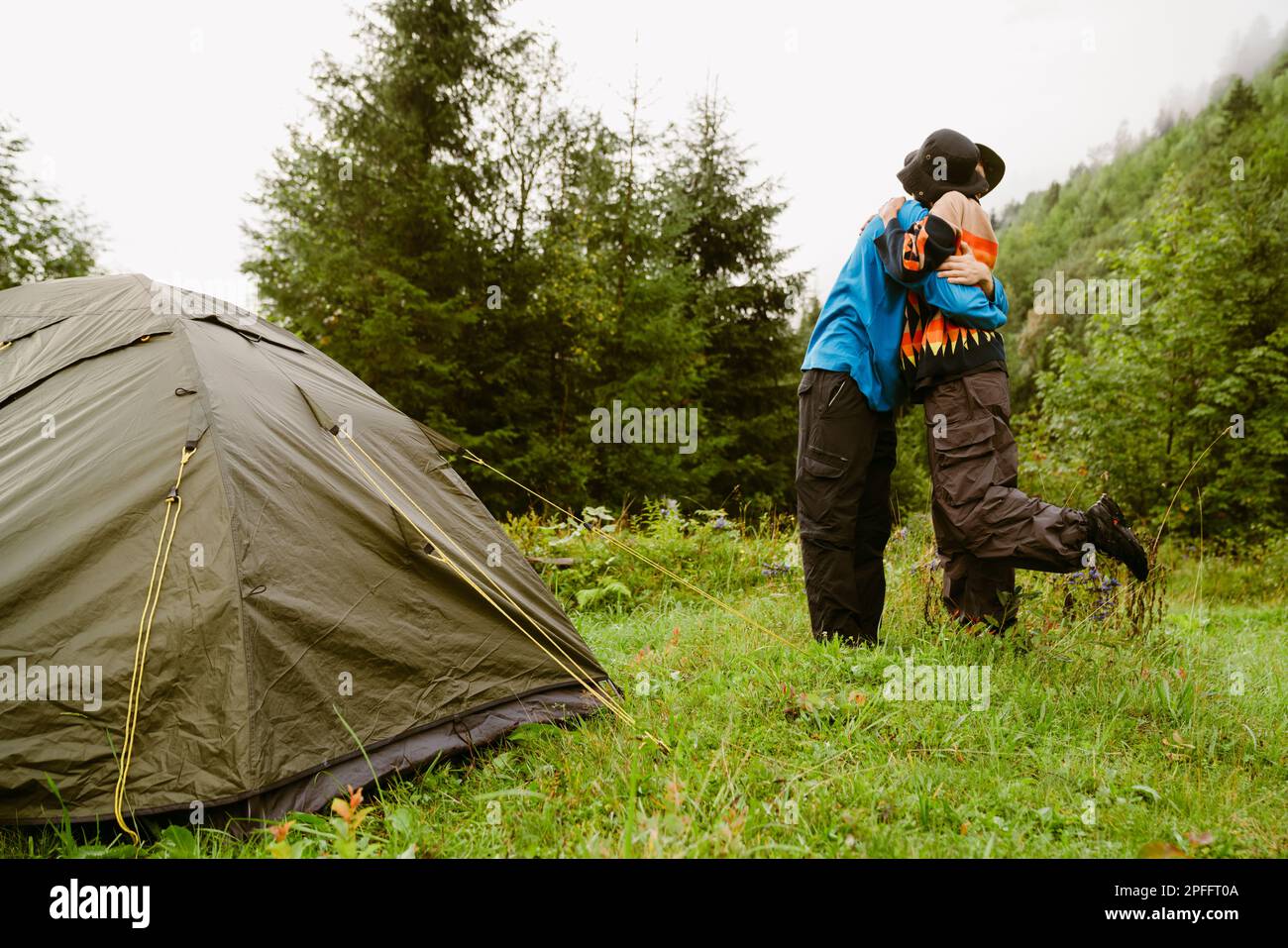 Young couple of travellers wearing panama hats hugging while standing near camping tent in green forest Stock Photo