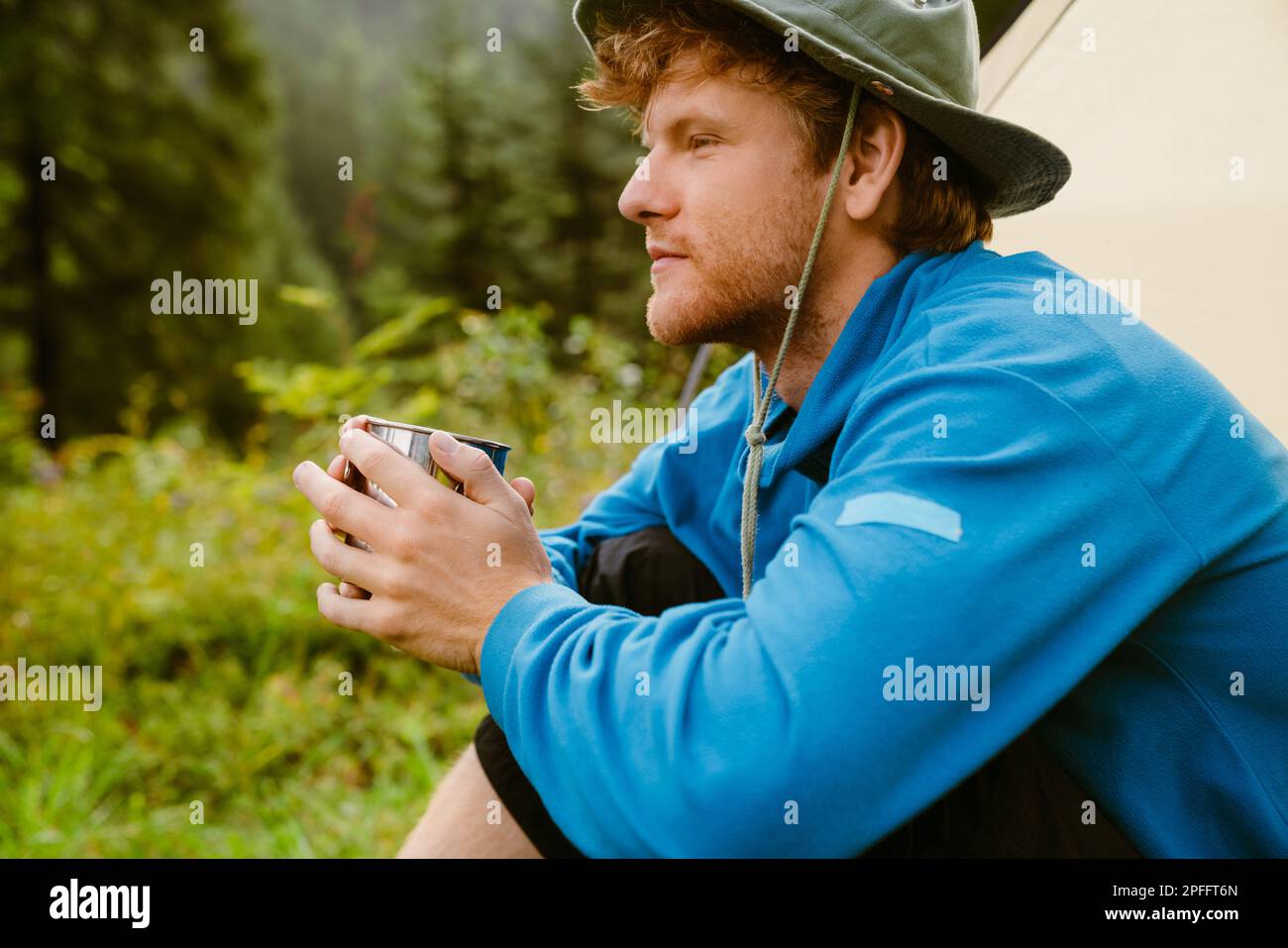 Young white tourist man drinking tea from metal cup while sitting at tent in forest Stock Photo