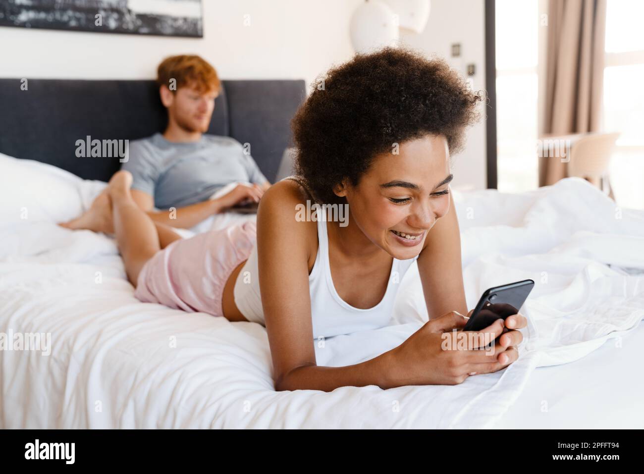 Young beautiful african curly smiling woman propping her head , holding and using her phone, while lying with her boyfriend who working with laptop on Stock Photo