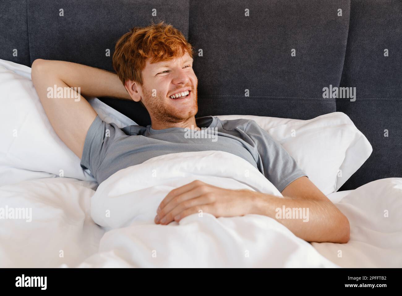 Young handsome smiling redhead man in gray t-shirt with hand behind his head looking aside lying on bed at home Stock Photo