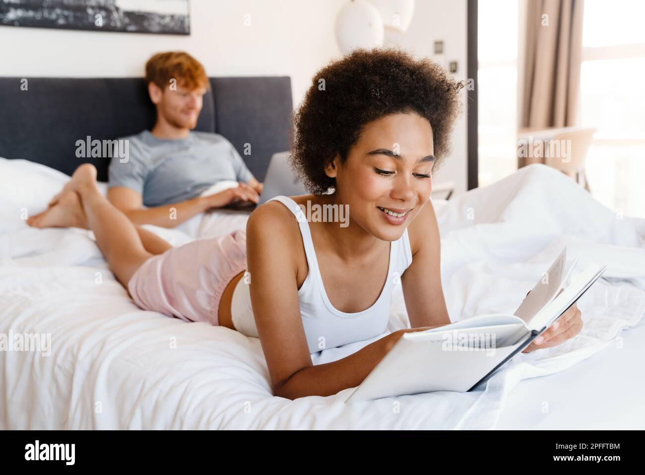 Young beautiful african curly smiling woman reading book lying with her boyfriend who working with laptop on bed at home Stock Photo