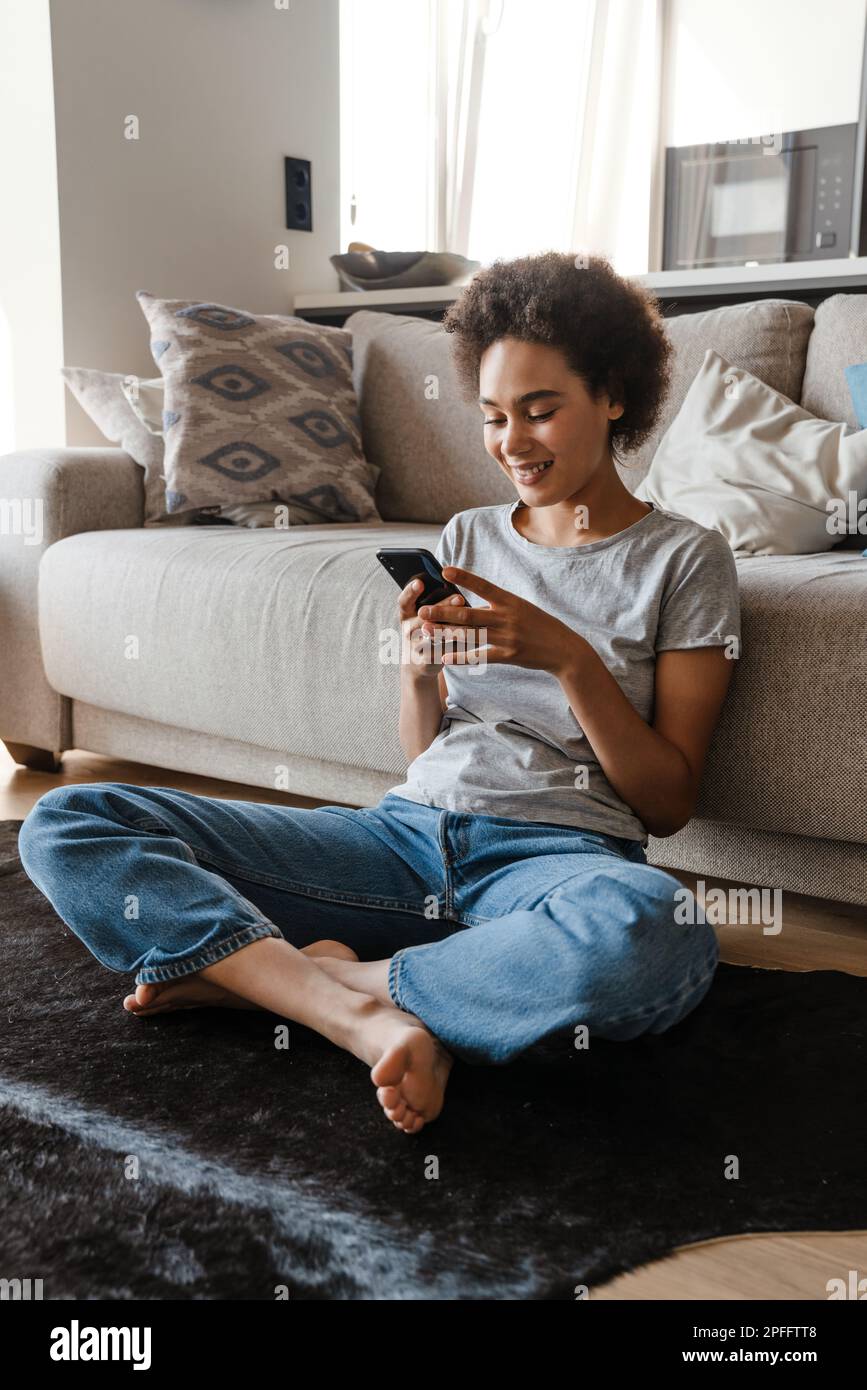 Young beautiful smiling curly african woman holding and using her phone sitting on carpet on the floor in lotus pose leaning on couch in cozy living r Stock Photo