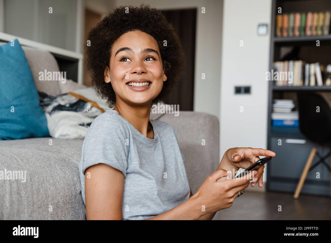 Portrait of young beautiful smiling curly african woman holding phone and looking aside ,sitting on the floor leaning on couch at home Stock Photo