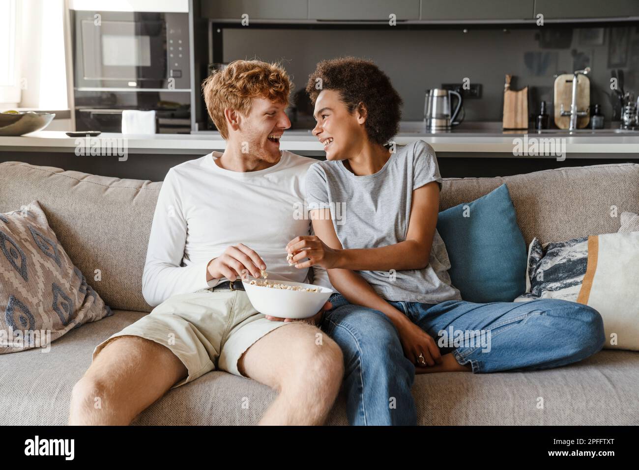 Young beautiful happy interracial couple eating popcorn and looking at each other, while sitting on couch in cozy sunny living room at home Stock Photo