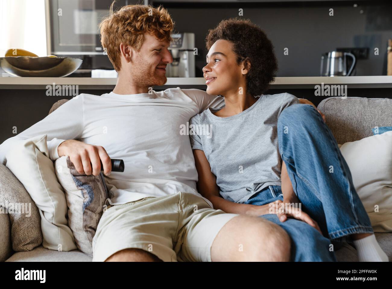 Young beautiful happy interracial couple hugging and looking at each other , while sitting on couch at home together. Handsome redhead man and beautif Stock Photo