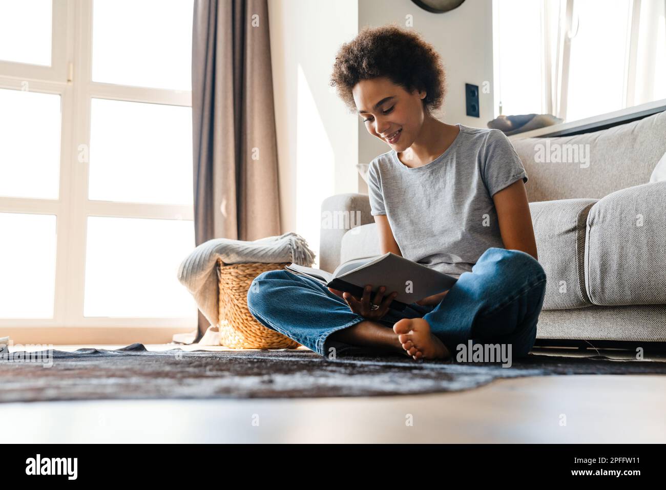 Young curly woman wearing jeans reading book while sitting on floor at home Stock Photo