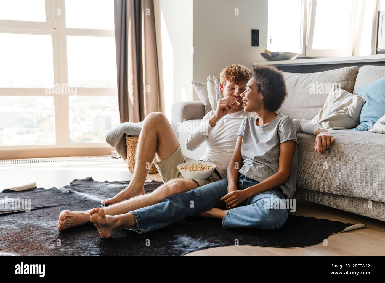 Young handsome redhead guy feeding his african girlfriend popcorn, while they sitting on the floor leaning on couch in cozy sunny living room at home Stock Photo