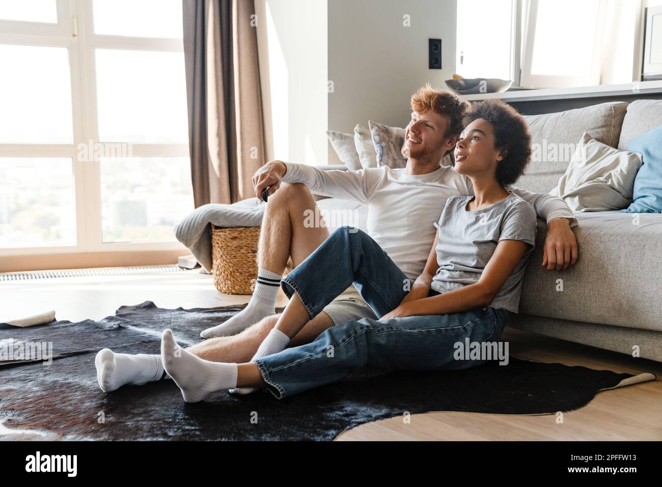 Young beautiful happy smiling interracial couple watching tv , while sitting on the floor leaning on couch at home together. Handsome redhead man and Stock Photo