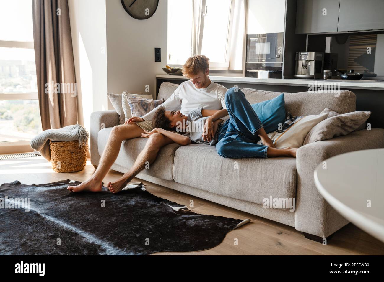Young cheerful multinational couple hugging while resting on sofa at home Stock Photo