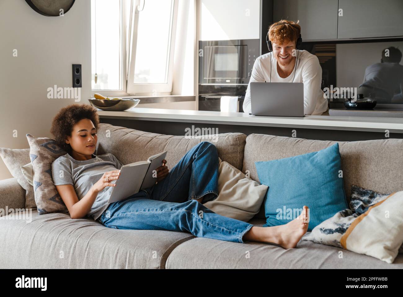 Young multinational couple using laptop and reading book while resting at home Stock Photo
