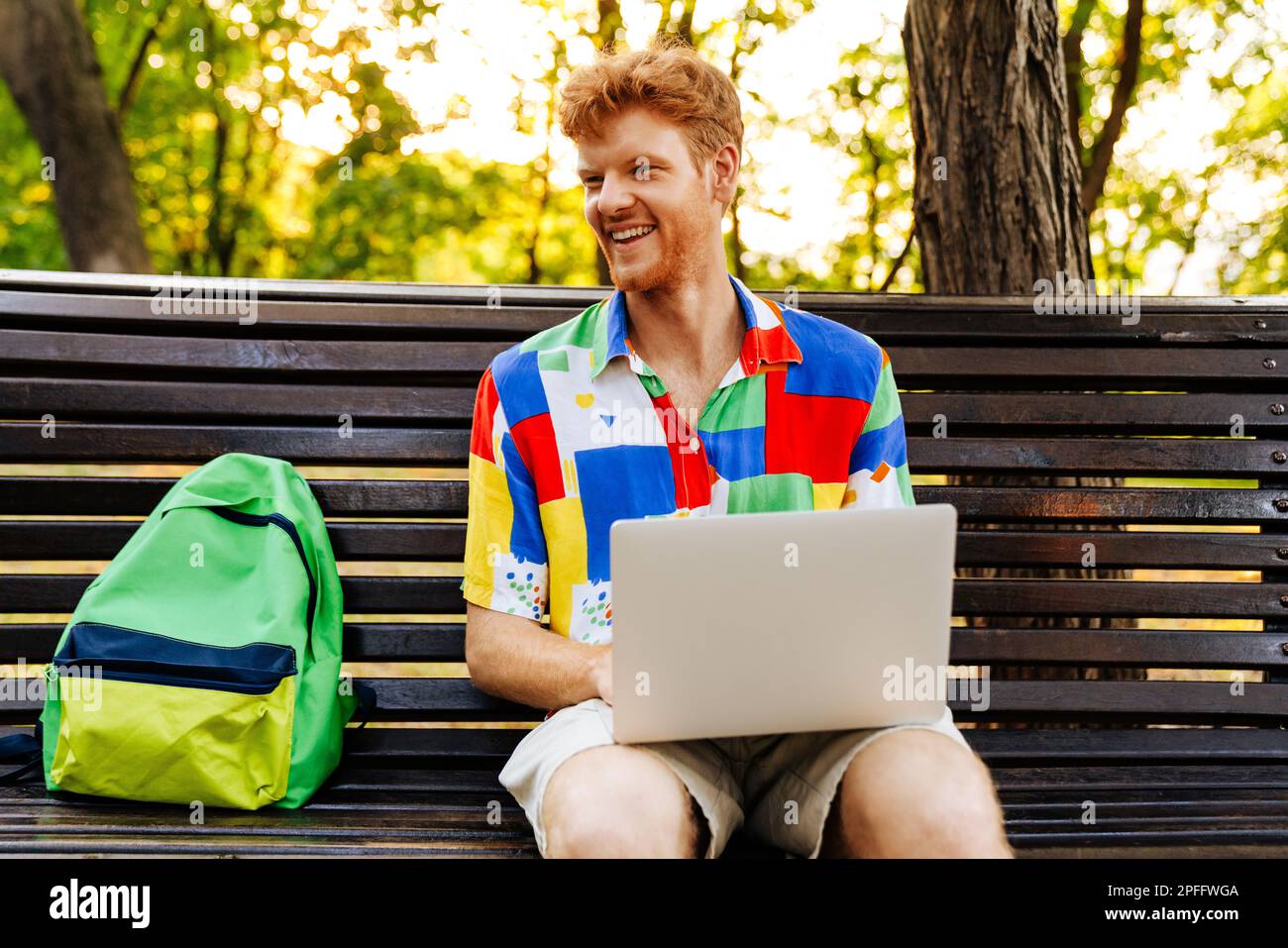 Young handsome smiling redhead man with laptop looking aside, while sitting with backpack on bench in park Stock Photo