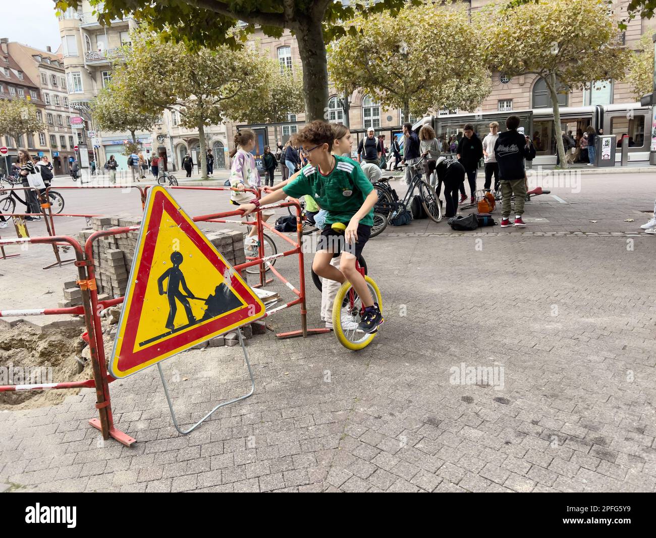 Strasbourg, France - Sep 23, 2022: large group of people gather around a working man fixing bicycles and unicycles on the side of the road. Trees line Stock Photo