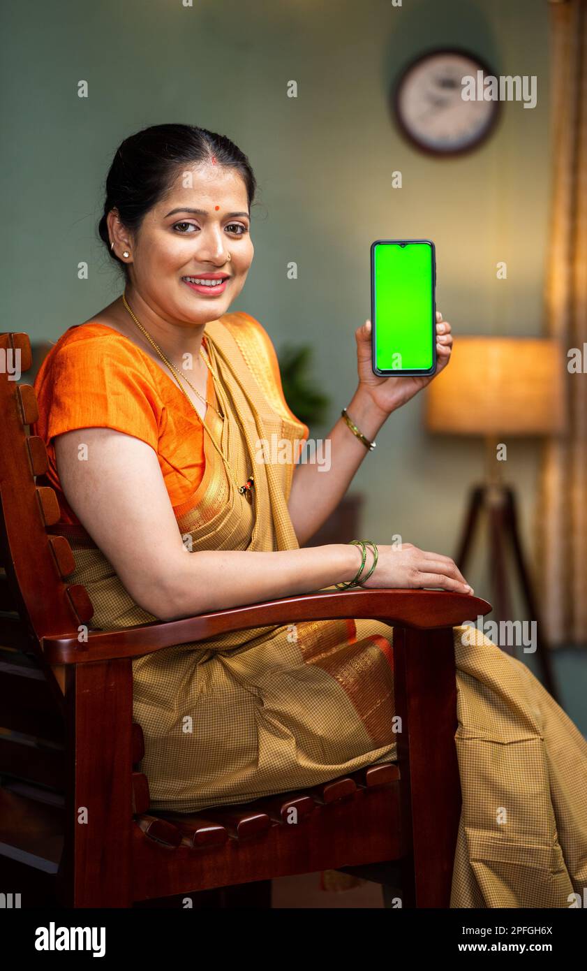 vertical shot of happy smiling woman showing green screen mobile phone by looking at camera on chair - concept app or application promotion Stock Photo