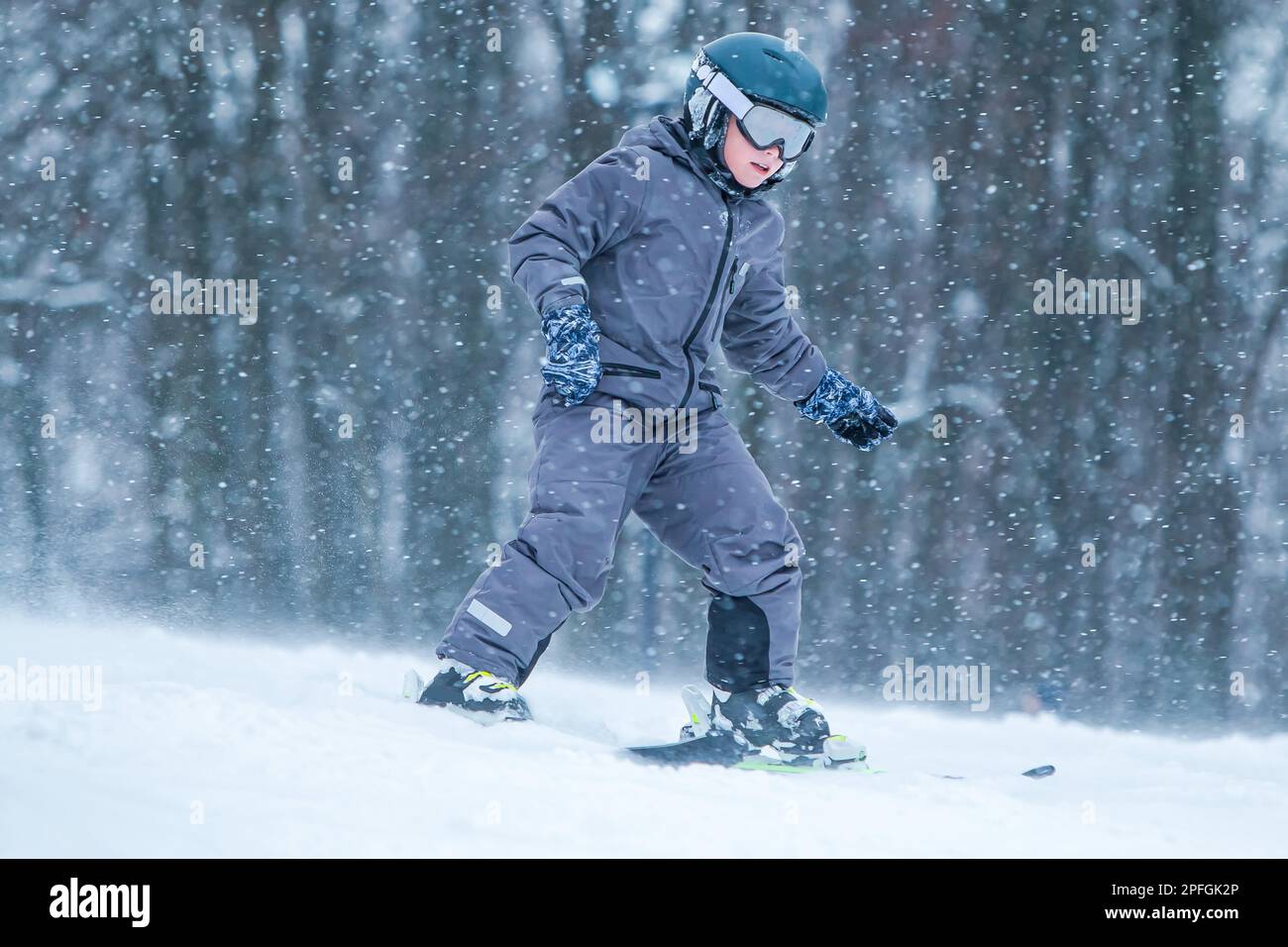 Little skier riding downhill with snow splash. Child skiing in mountains. Active teenage boy with safety helmet, goggles and ski poles running down Stock Photo