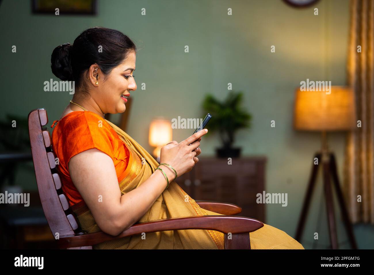 Middle woman in sare buy using mobile phone on chair at home - concept of technology, social media sharing, internet. Stock Photo