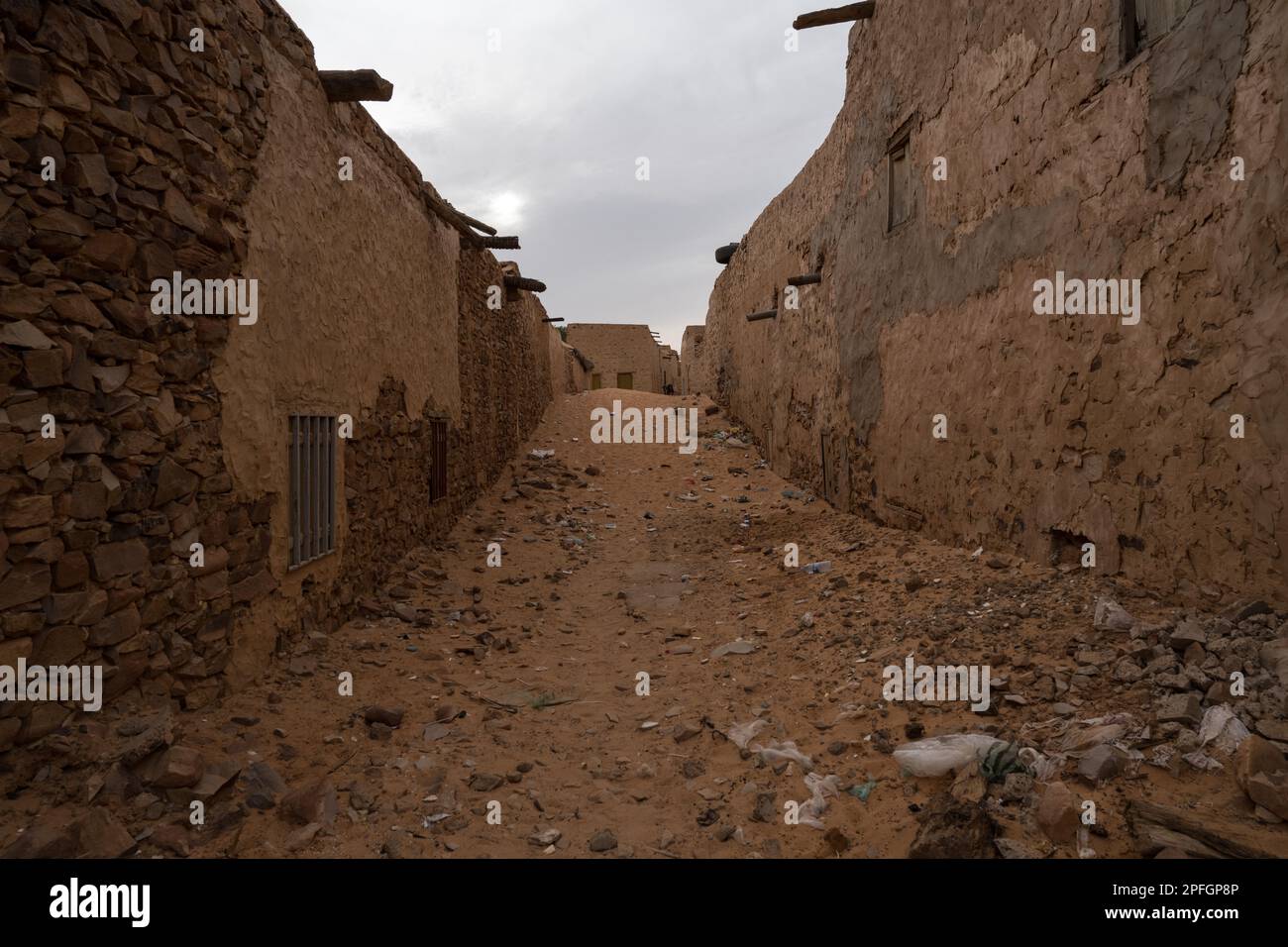 A street in the ancient city of Chinguetti, Mauritania, filled with sand and flanked by historic stone buildings, with goats standing in foreground. Stock Photo