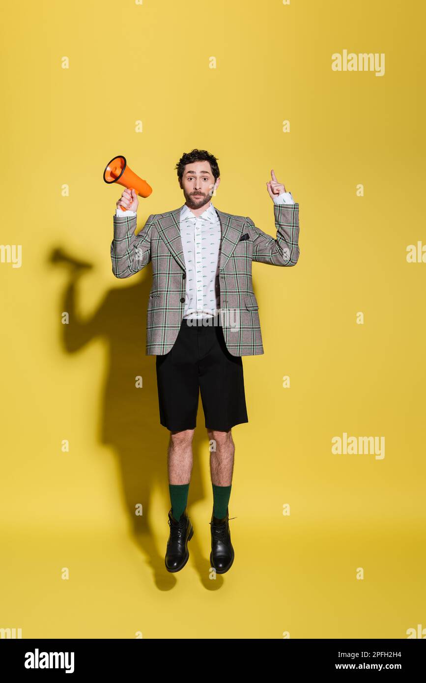 Fashionable man pointing with finger and holding loudspeaker while jumping on yellow background,stock image Stock Photo