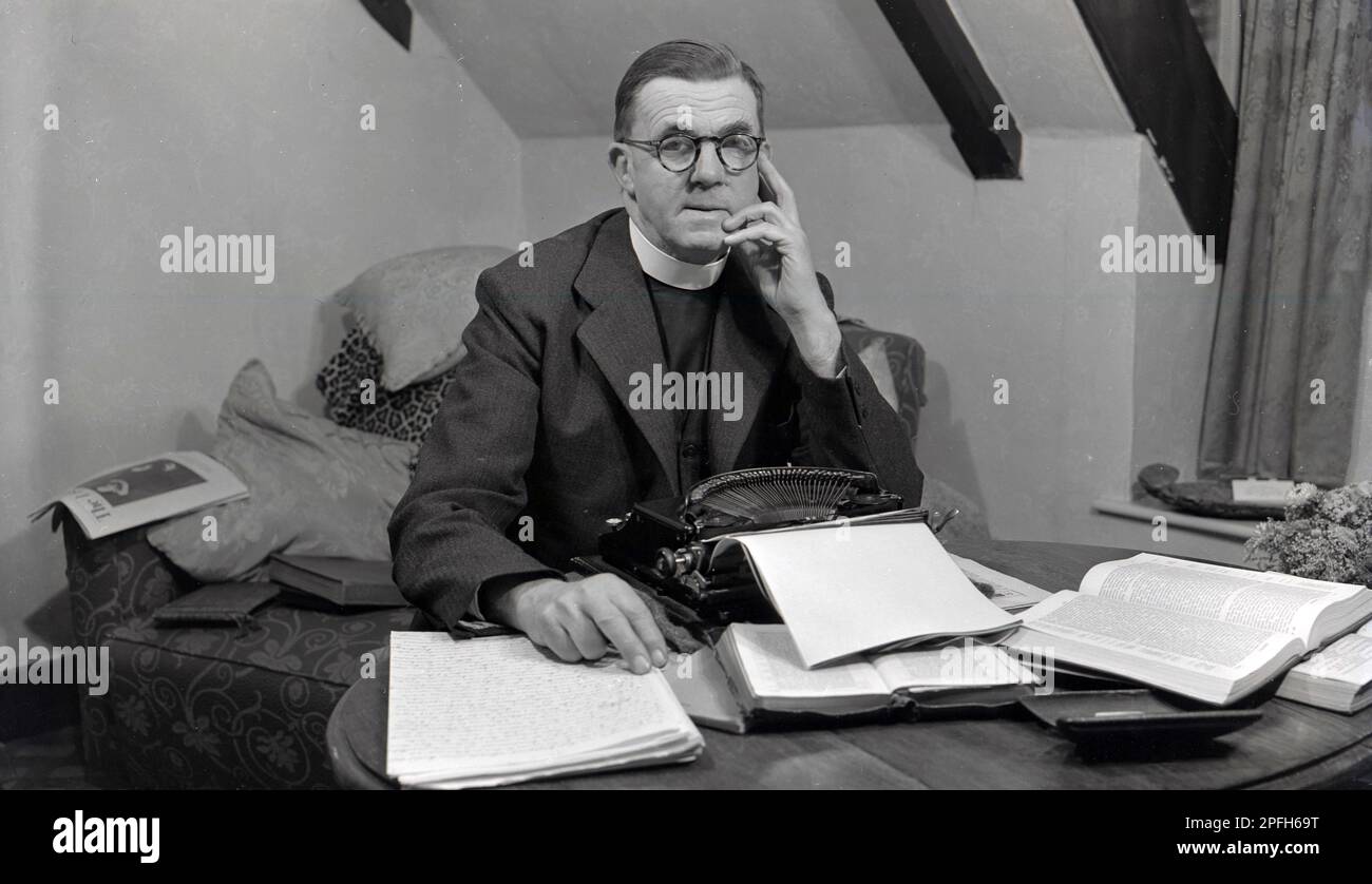 1940s, historial, a vicar, wearing a clerical collar, stting inside at a desk at a typewriter of the era, wiith paper in and surrounded by prayer bookd and possibly the bible, USA. Stock Photo