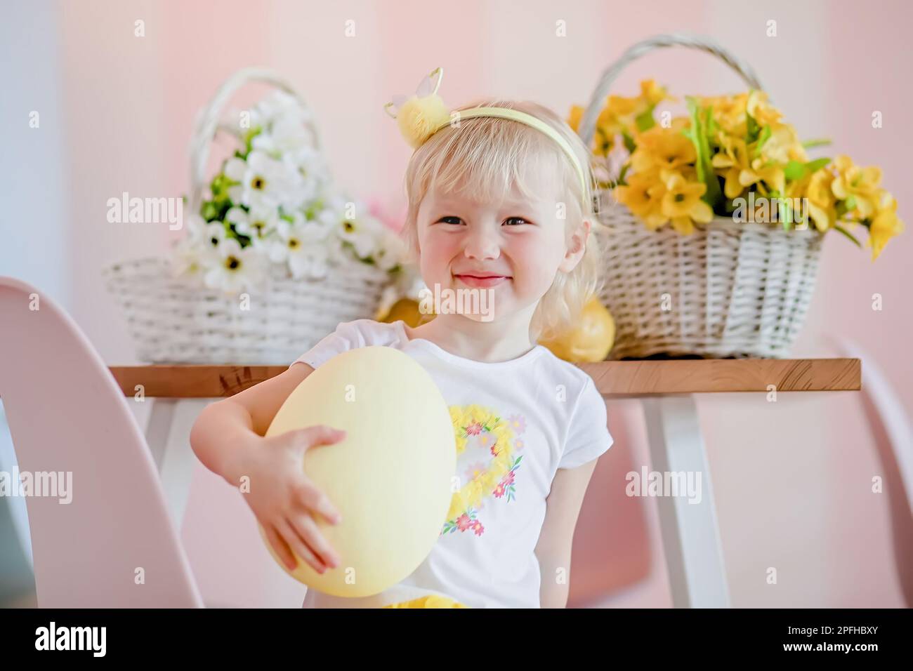 Easter egg hunt at home. Adorable happy little blonde girl with short hair in a white dress holding a huge painted Easter egg. Stock Photo