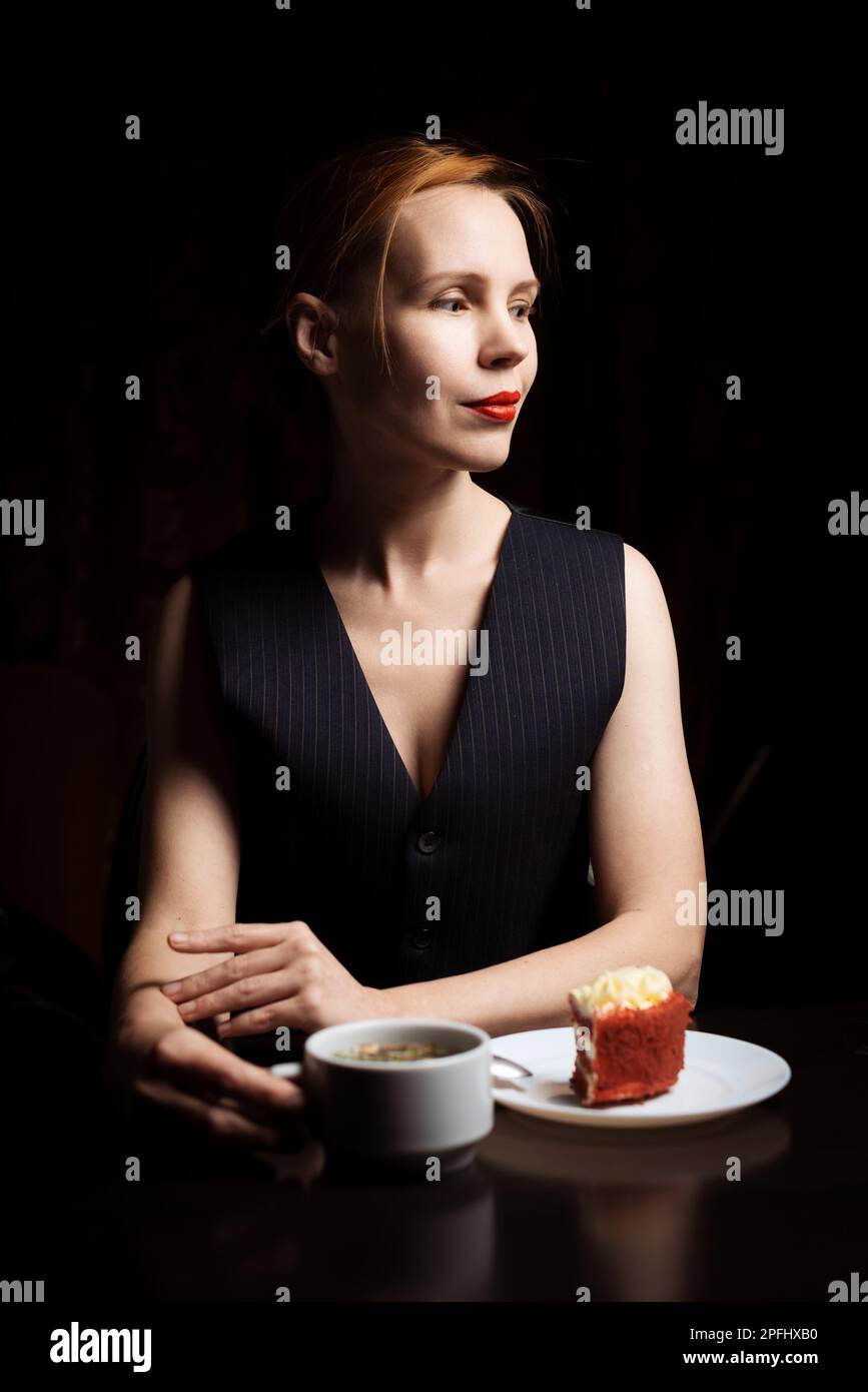 Woman at the table with tea and cake in the dark. Attractive forty-year-old woman in a black vest. Tea drinking. Loneliness. Stock Photo