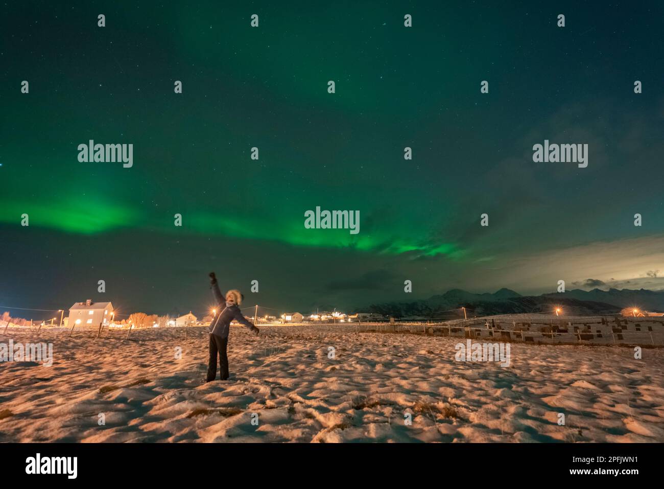 wunderbare Nordlichter über dem Dorf Hillesøy. tanzende Frau auf Feld unter der tanzenden Aurora Borealis. Hell erleuchtete Häuser und Straßenlaternen Stock Photo