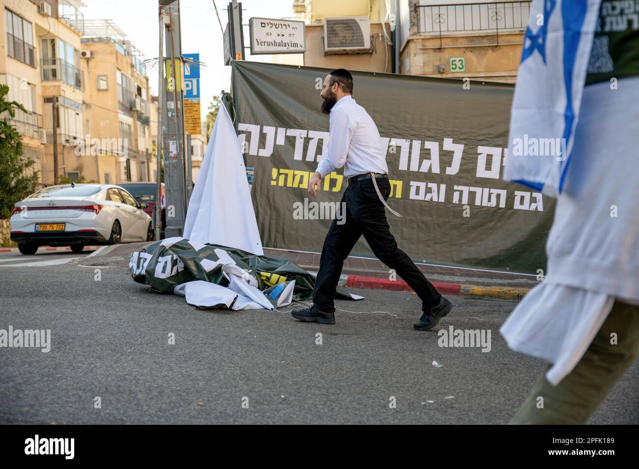 Bnei Brak, Israel. 16th Mar, 2023. An orthodox man passes next to a protest against the reform banner that says 'Haredim/anxious for the fate of the country, Torah as well as martial theory/Torah' during the demonstration. The soldiers opened a “recruiting centre” to IDF military service in a protestation act against the judicial overhaul in the Ultra-Orthodox city of Bnei Brak. Credit: SOPA Images Limited/Alamy Live News Stock Photo