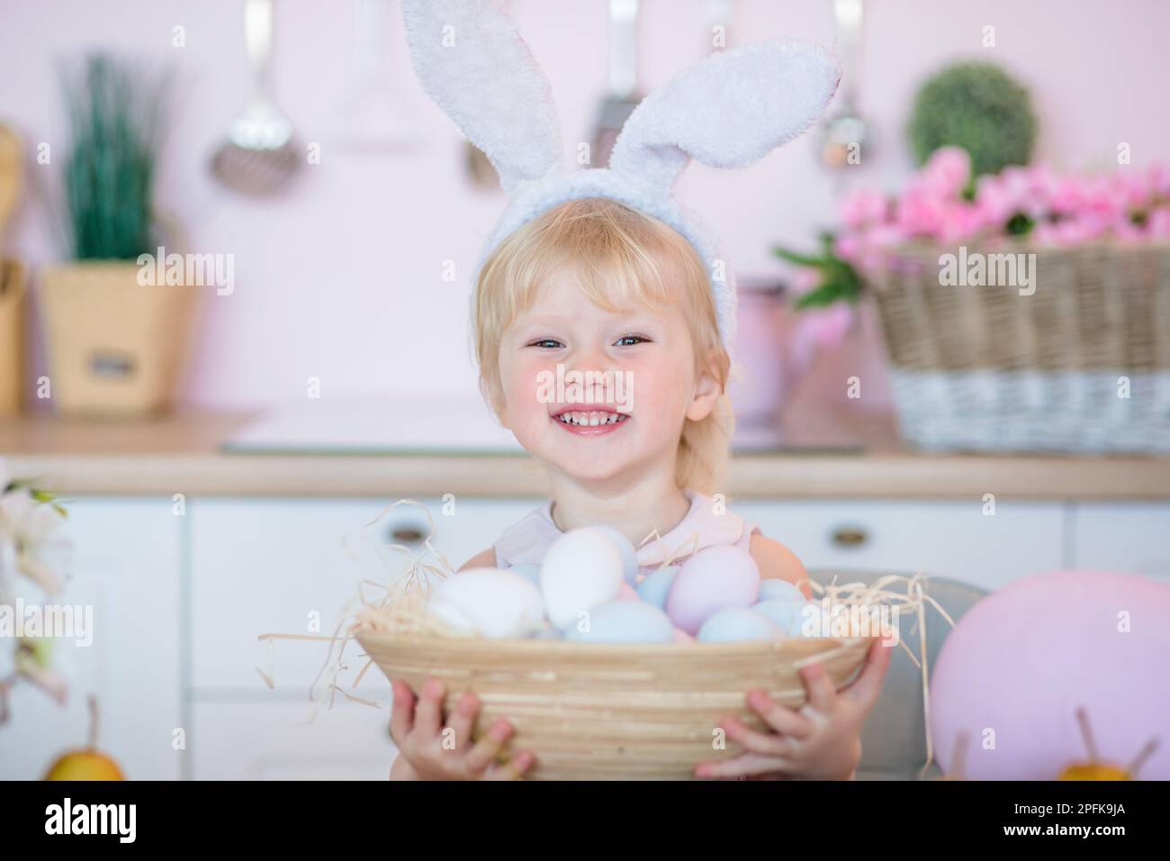 Little girl holds a basket with painted Easter eggs. Happy child in the bright kitchen at home on Easter. Stock Photo