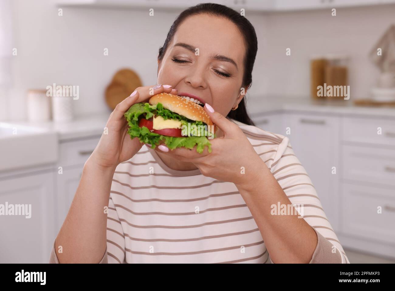 Overweight woman eating burger in kitchen at home Stock Photo