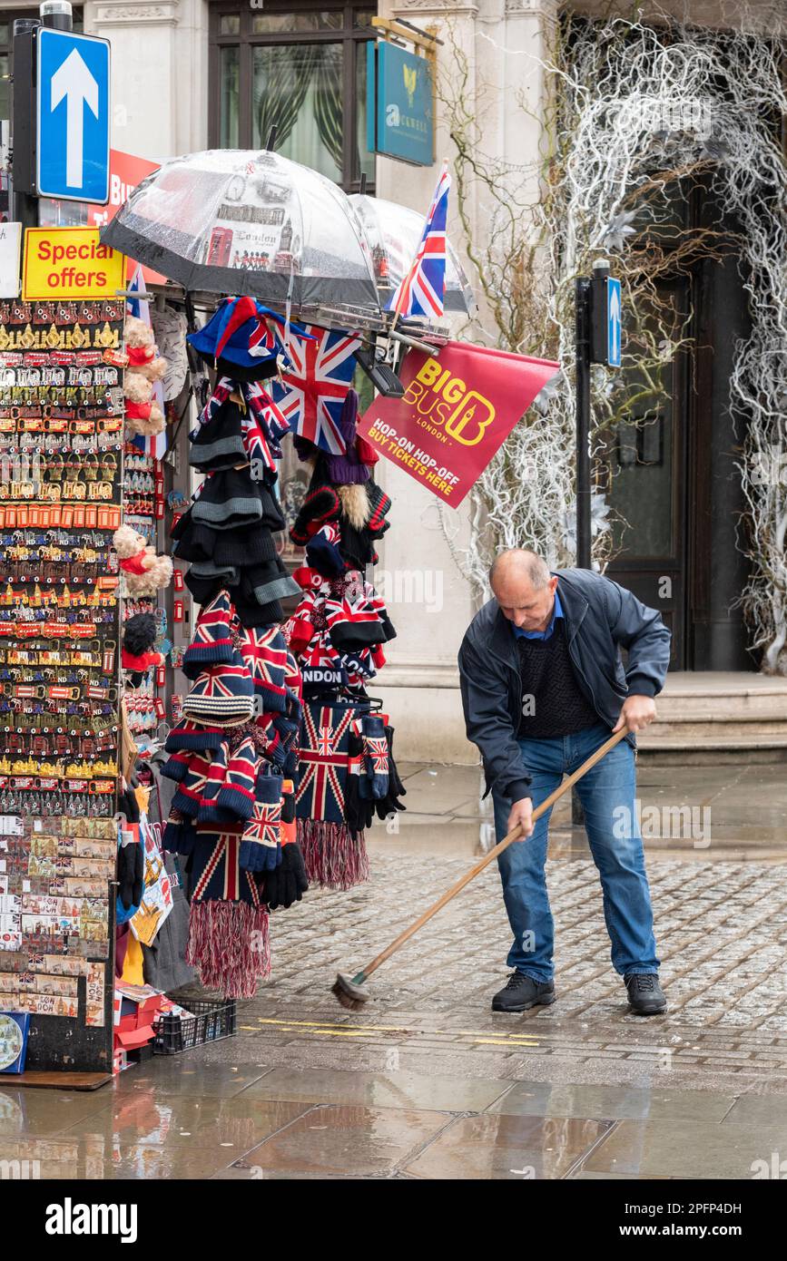 Westminster, London, UK. 18th Mar, 2023. The day has begun wet and overcast. Souvenir vendor sweeping rainwater away from sales stand Stock Photo