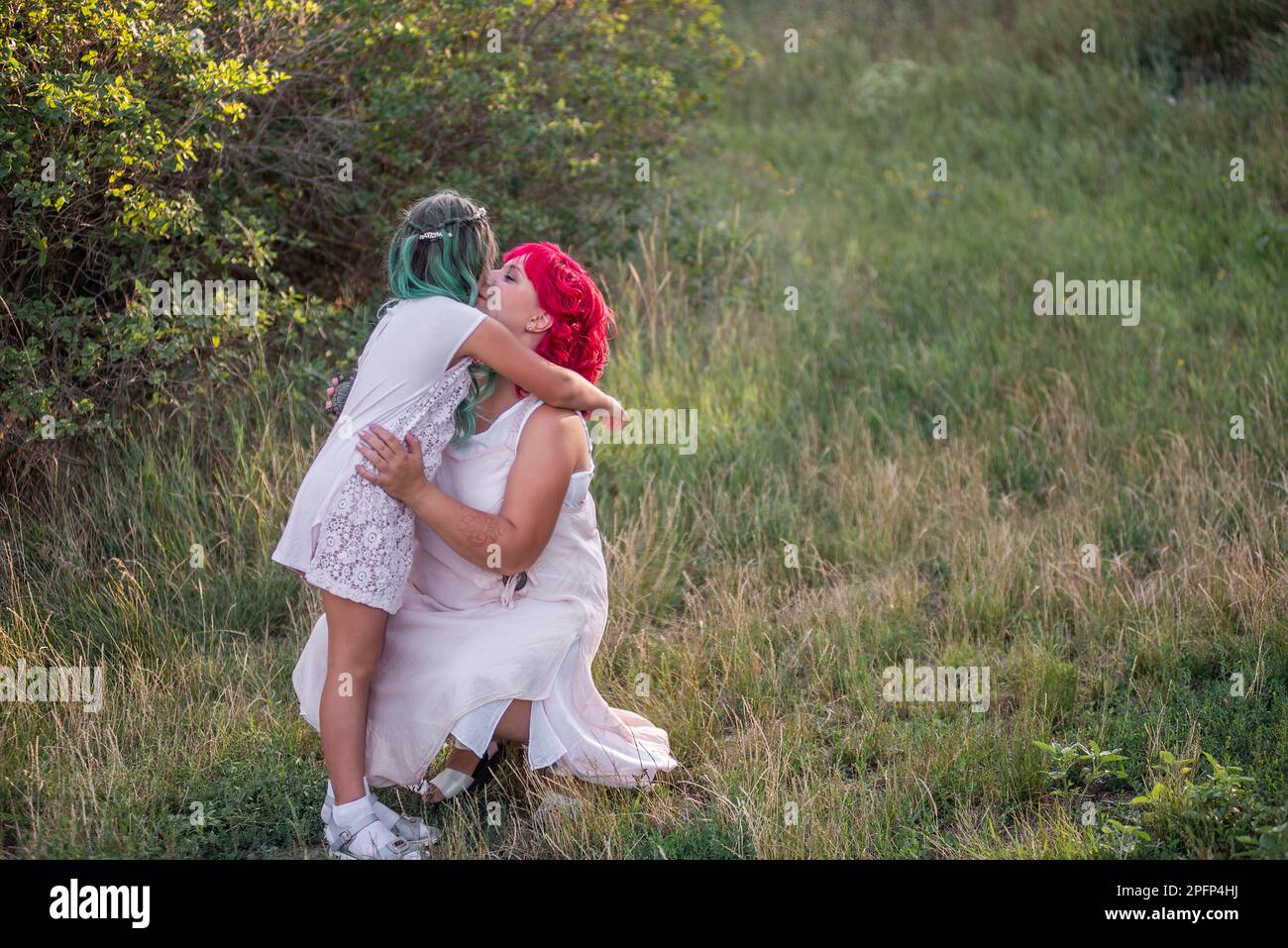 Bright, diversity, extraordinary mother and daughter hugging in the forest. Woman has pink hair, the girl has green hair. Tender motherhood, happy chi Stock Photo