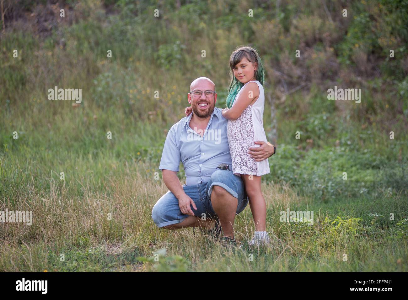 Bald father in glasses hugs daughter with green hair in nature. Diversity family portrait smilling. Fathers Day. Care, child care. Children Protection Stock Photo