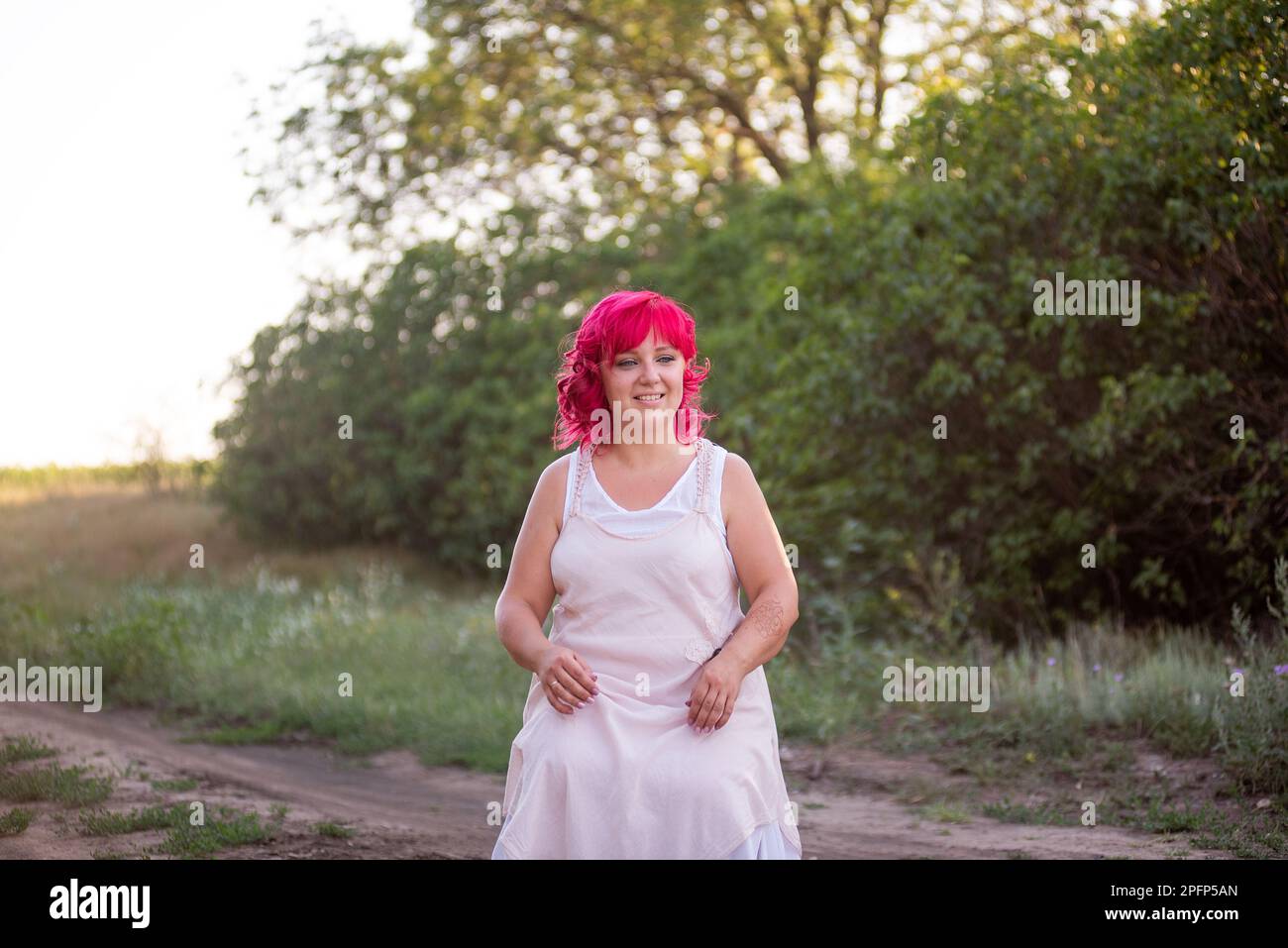 Bright diversity woman with pink hair portrait. The girl squatted down on a forest path into the sunset. Childfree girl, weekend trip. Individual, bol Stock Photo