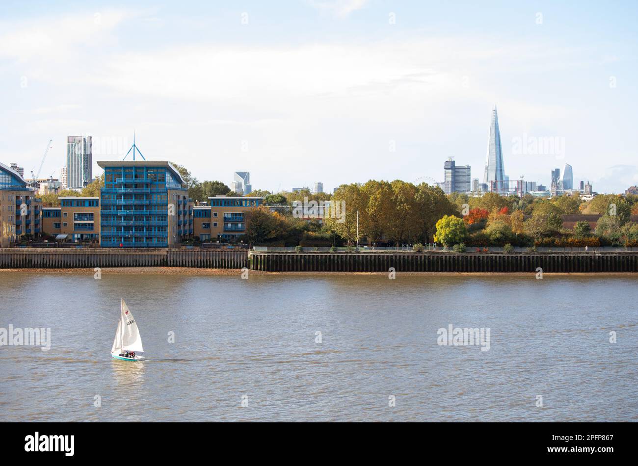 A small yacht sails on the Thames past the autumn London skyline including the Shard and the London Eye. Also past desirable riverside apartments. Stock Photo