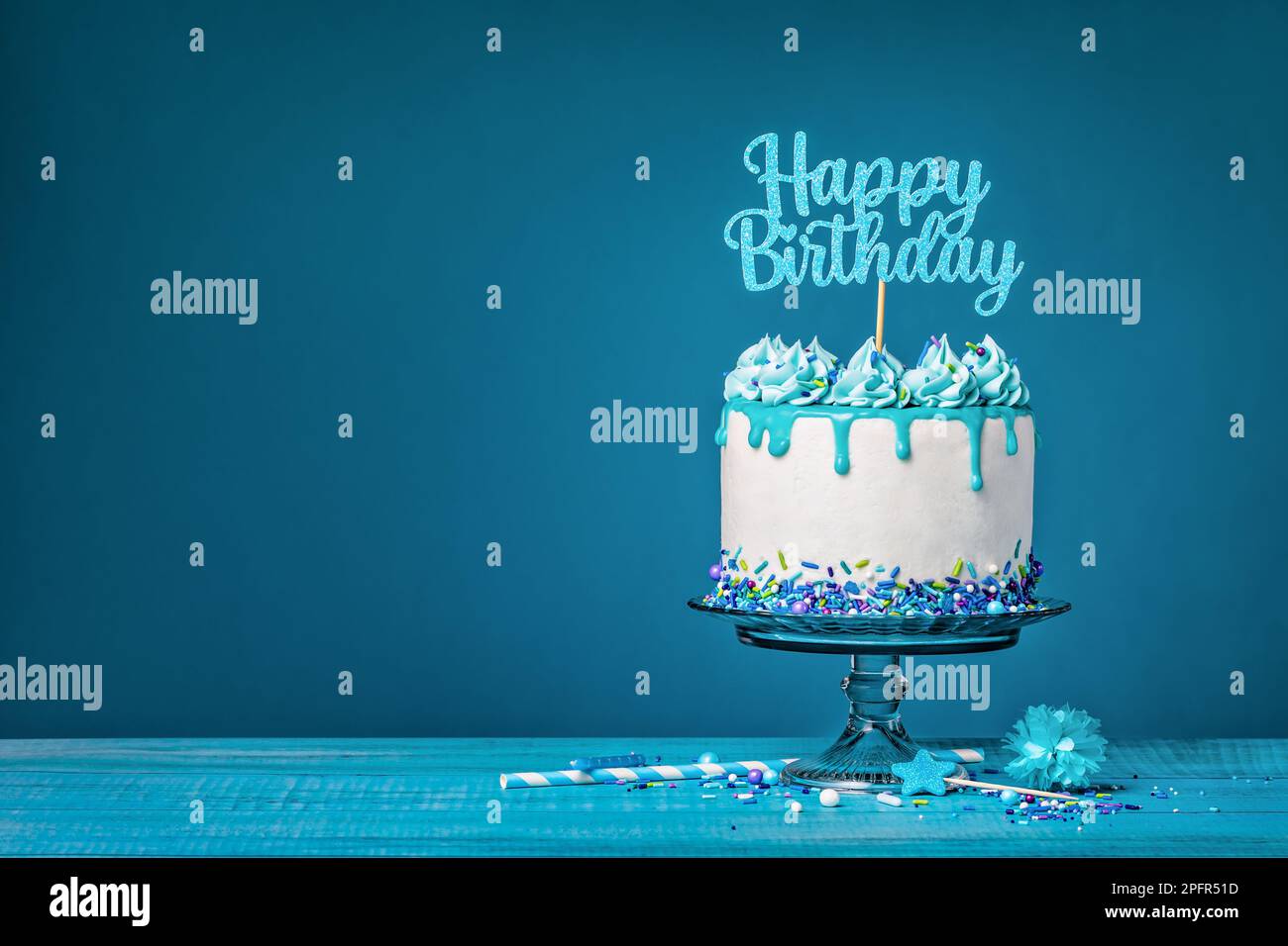 White drip cake with teal ganache, sprinkles, and a happy birthday topper over a blue background. Simple and trendy. Copy space. Stock Photo