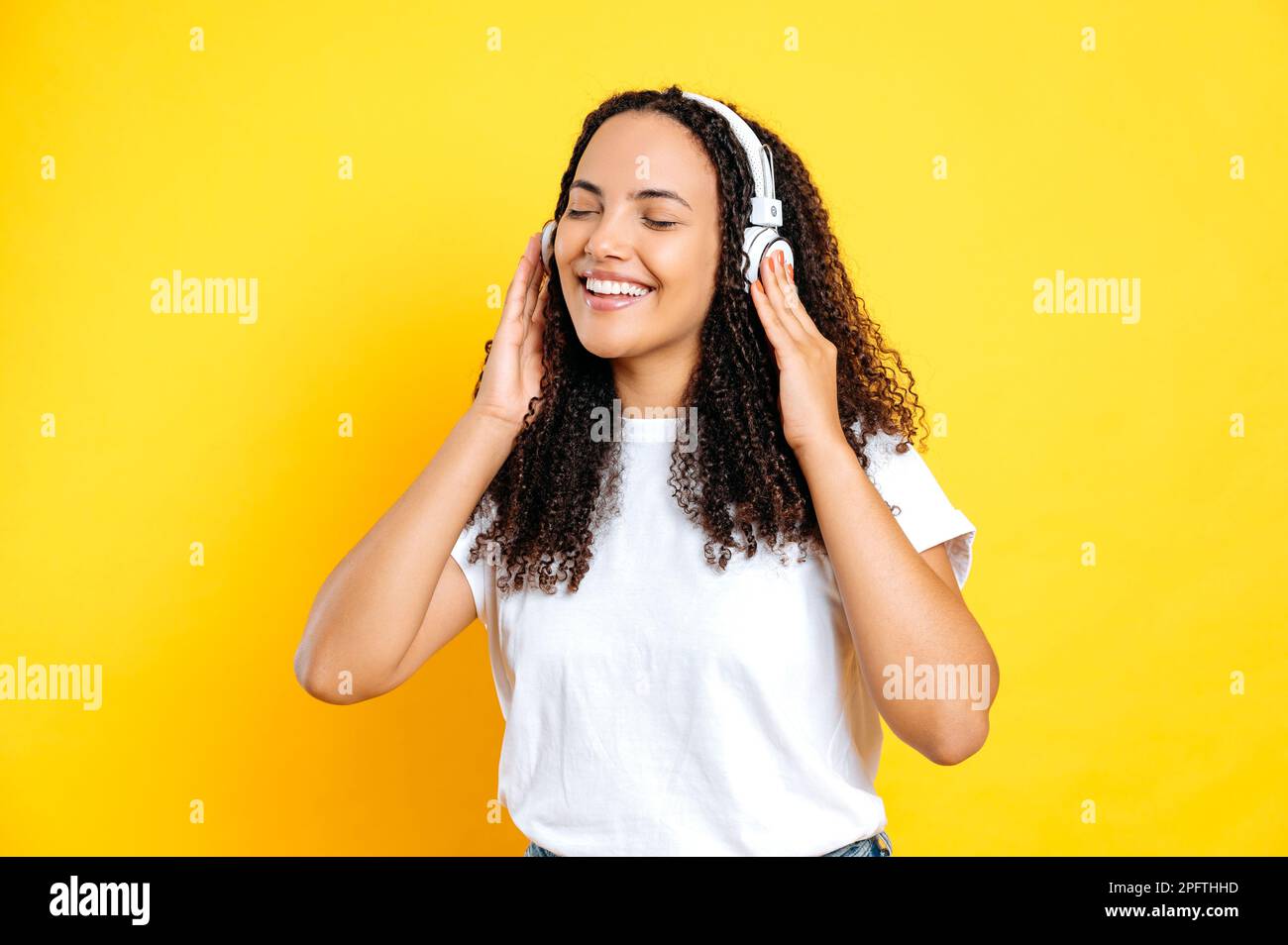Cheerful pretty brunette brazilian woman, in casual stylish wear, with headphones, enjoy her favorite music, dancing with eyes closed, relaxing, having fun on isolated yellow background, smiling Stock Photo