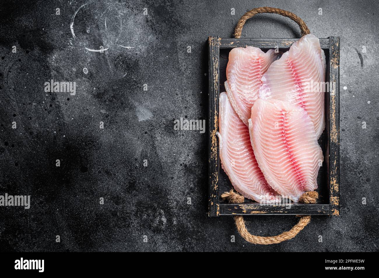 Fresh tilapia white fish fillet in a wooden tray. Black background. Top