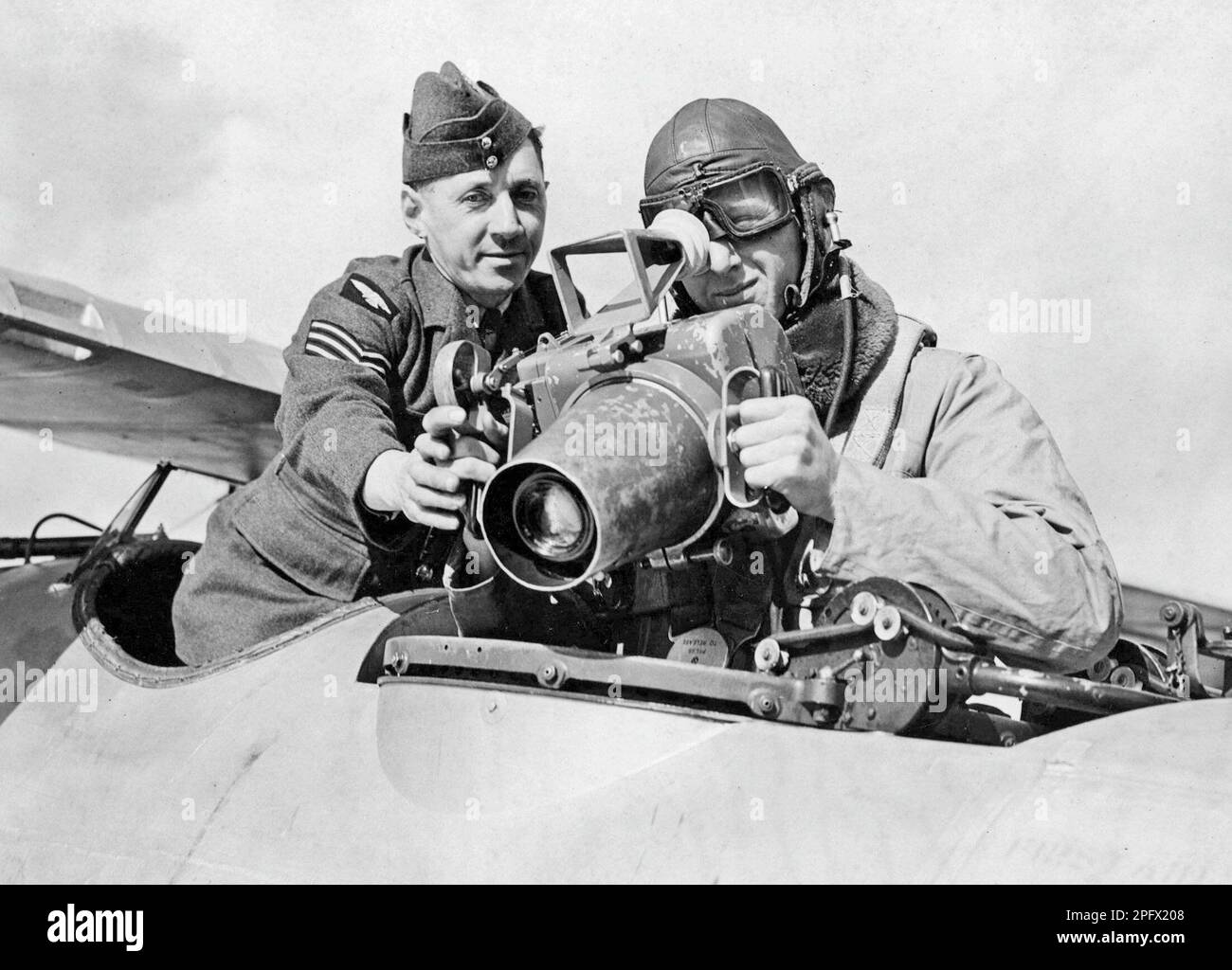 In the 1930s. A crewmember of a military airplane with a soldier at a camera mounted on the airplane. The camera might be new and some guidence in how to operate it is needed. The man dressed in a flying suit is using the sight and focus on something. England 1939 Stock Photo