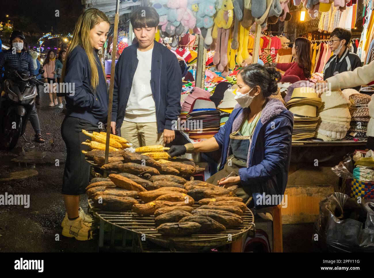 A young Vietnamese couple stop to buy grilled corn-on-the-cob at the bustling night market in Dalat, Vietnam. Stock Photo