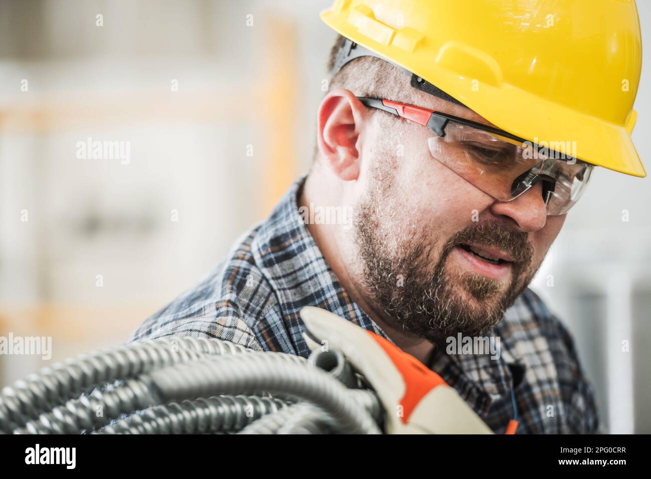Caucasian Professional Electric Contractor Worker in His 30s with Electric Conduit. Wearing Yellow Hard Hat and Protection Glasses. Stock Photo