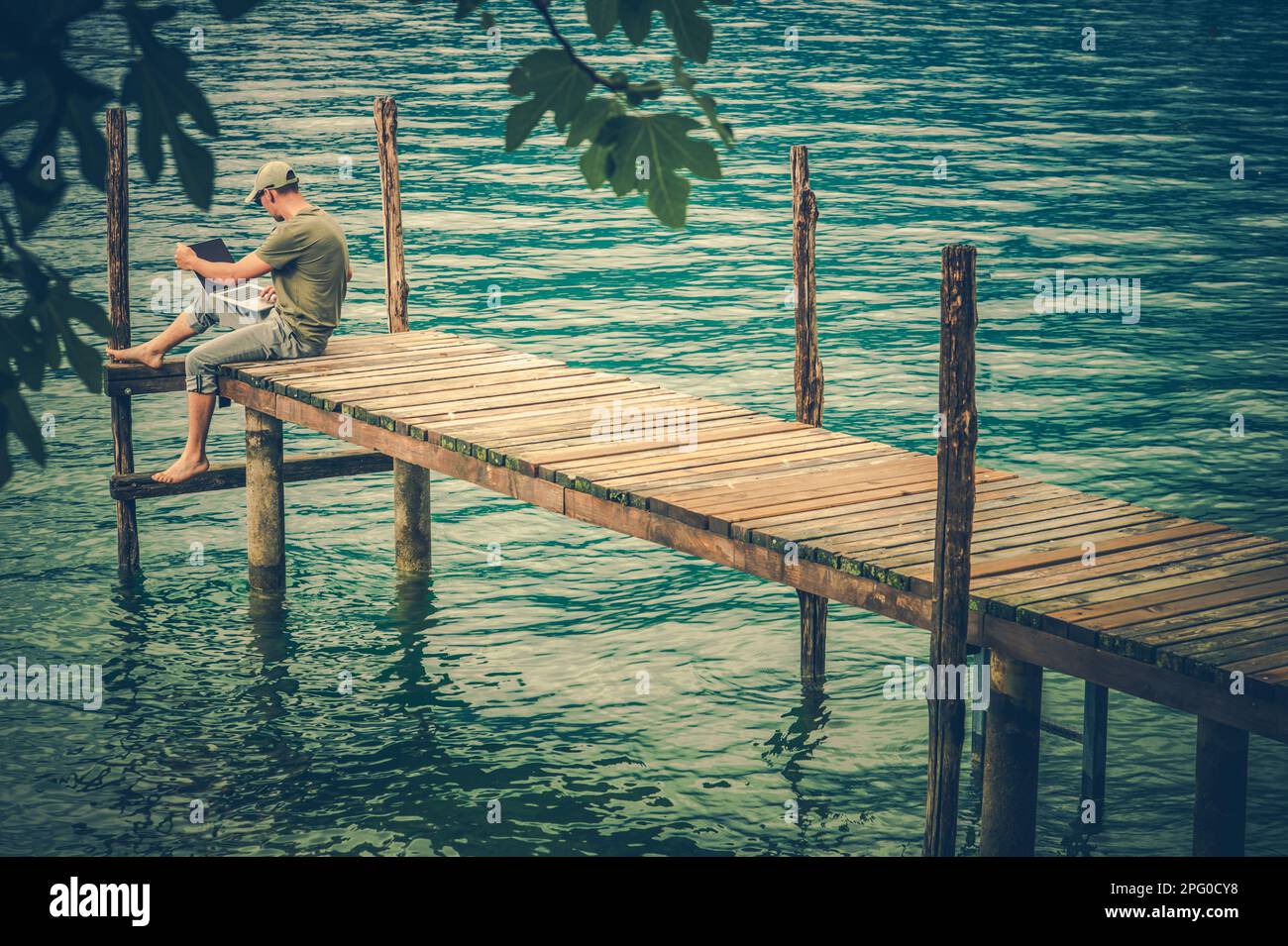 Computer Remote Work While on Vacation. Caucasian Man Using His Computer in Front of Scenic Lake. Seating on Wooden Pier Deck. Stock Photo
