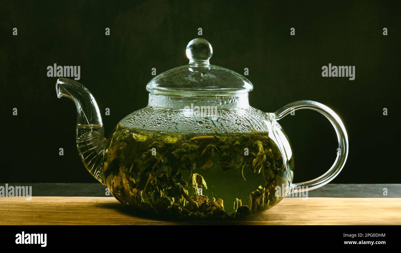 A glass teapot for brewing tea stands on a wooden board on a dark