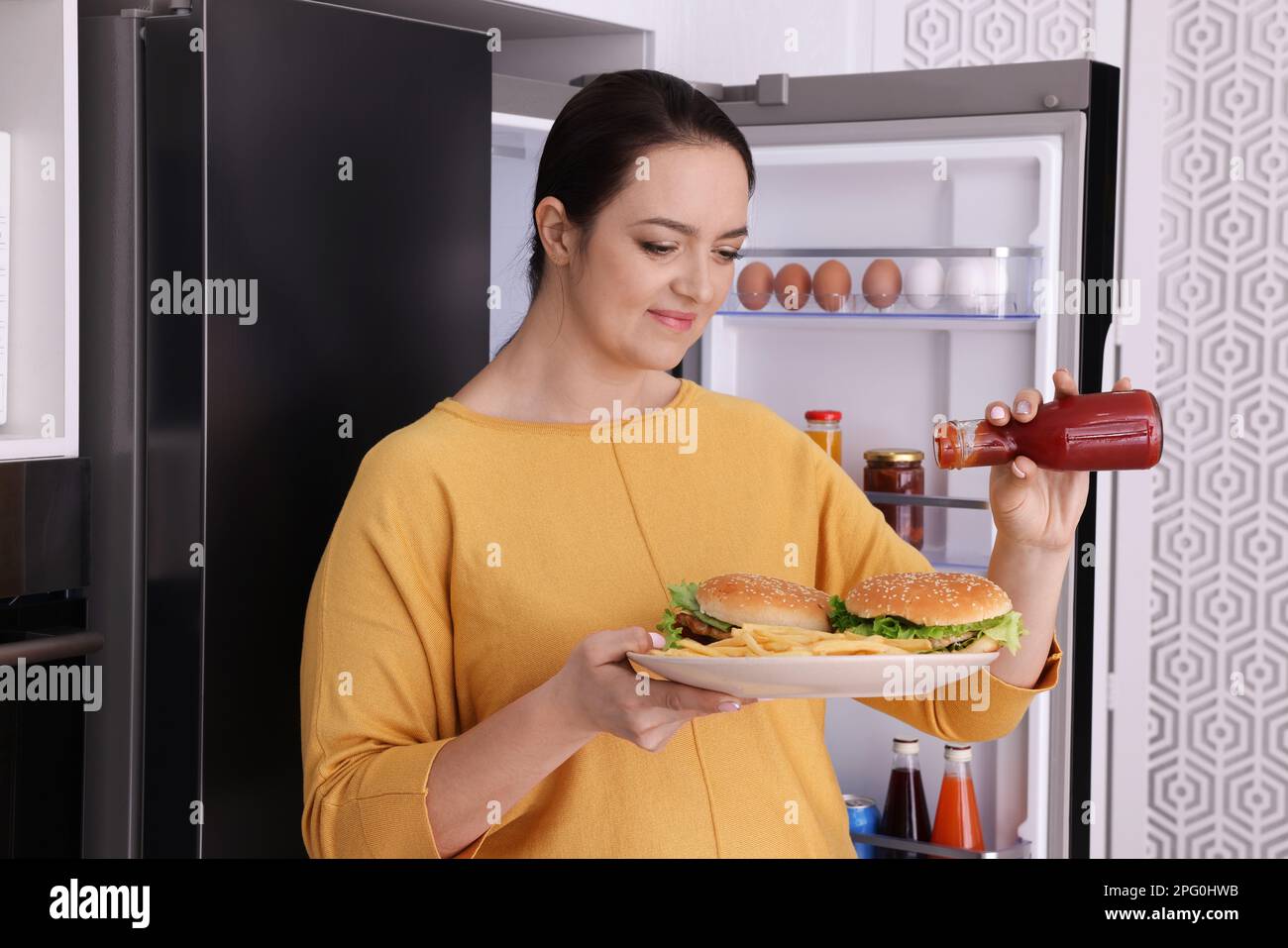 Overweight woman with ketchup and burgers near fridge in kitchen Stock Photo